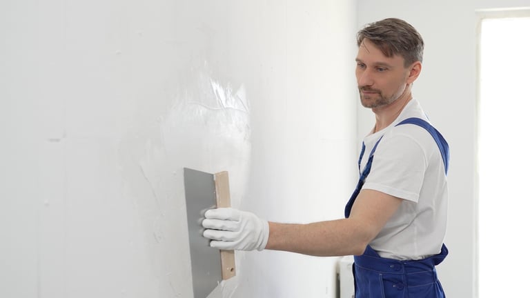 Male construction worker wearing protective gloves and blue construction coveralls, spreading plaster smoothly across wall using professional drywall taping knife during renovation project. Portrait view.