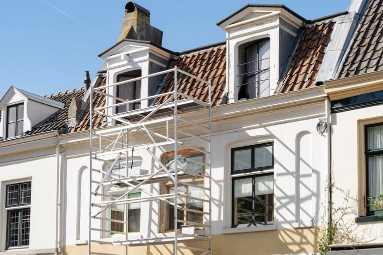 Scaffolding is erected against a white building with dormer windows and a red tile roof for maintenance.